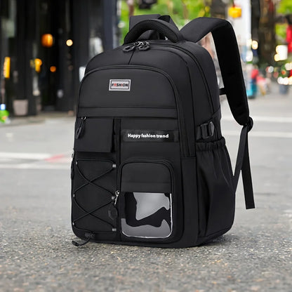 A black backpack standing upright on a city street. The backpack has multiple zippered compartments, side mesh pockets, a top handle, padded shoulder straps, and a small front patch with the brand name 'FISCHER'.
