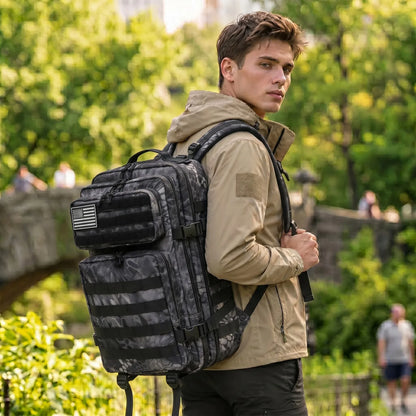 A man wearing a beige hooded jacket and black pants carries a large black tactical backpack with a camouflage pattern and multiple front pockets, standing outdoors in a park with green trees in the background.