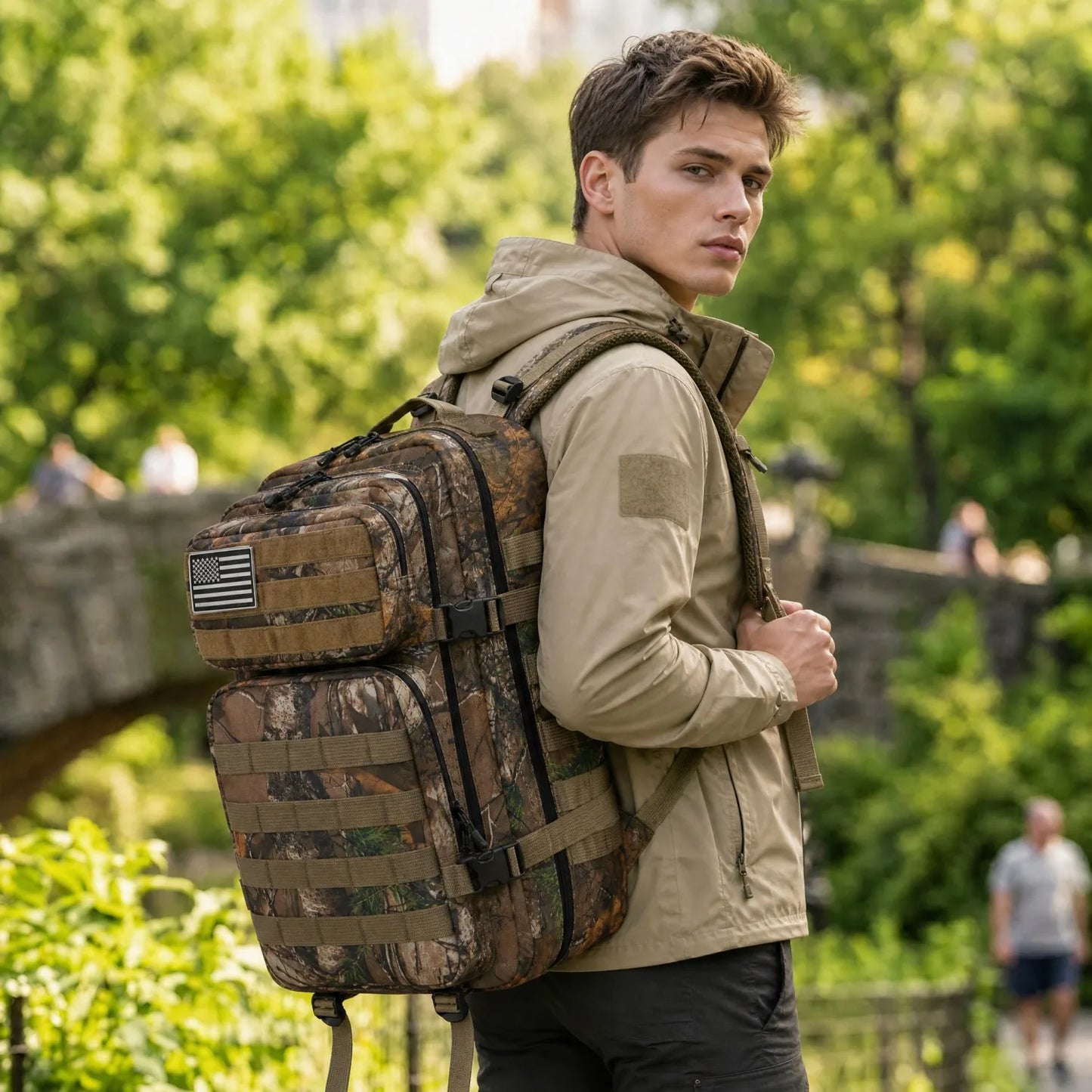 A man wearing a beige hooded jacket and black pants carries a large camouflage backpack with multiple straps and a small American flag patch on the side, standing outdoors in a park with green trees and stone bridges in the background.