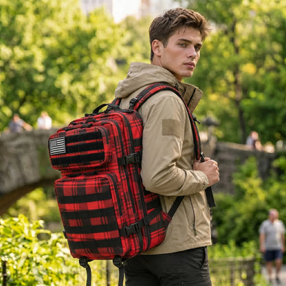 A man wearing a beige hooded jacket and black pants carries a large red and black plaid backpack with multiple compartments and a small American flag patch, standing outdoors in a park with green trees in the background.