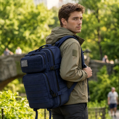 A man wearing a navy blue tactical backpack with a small American flag patch on the side, standing in a park with green trees in the background.