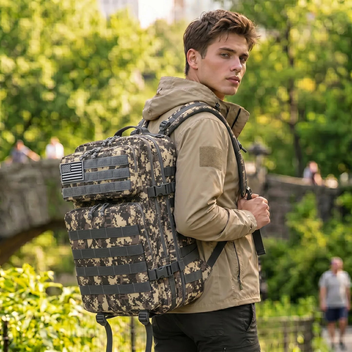 A man wearing a tan hooded jacket and black pants carries a large tactical backpack with a digital camouflage pattern in an outdoor park setting.