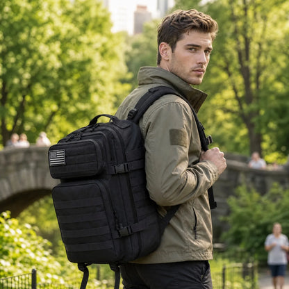 A man wearing an olive green jacket and black pants carries a large black tactical backpack with multiple compartments and a small American flag patch on the side, set against a blurred green park background.