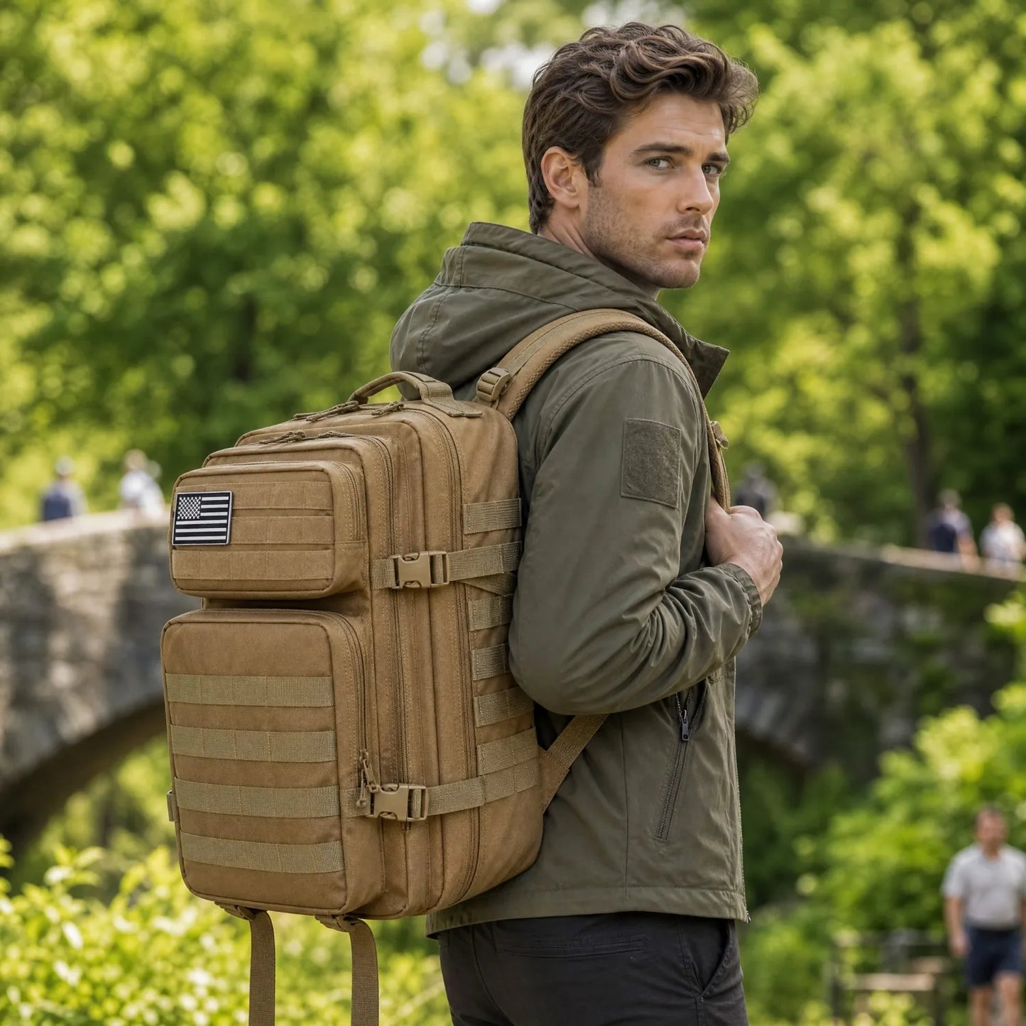 A man wearing an olive green jacket and black pants carries a large tan tactical backpack with MOLLE webbing and a small American flag patch on the side, photographed outdoors in a park-like setting with blurred green trees and a stone bridge in the background.