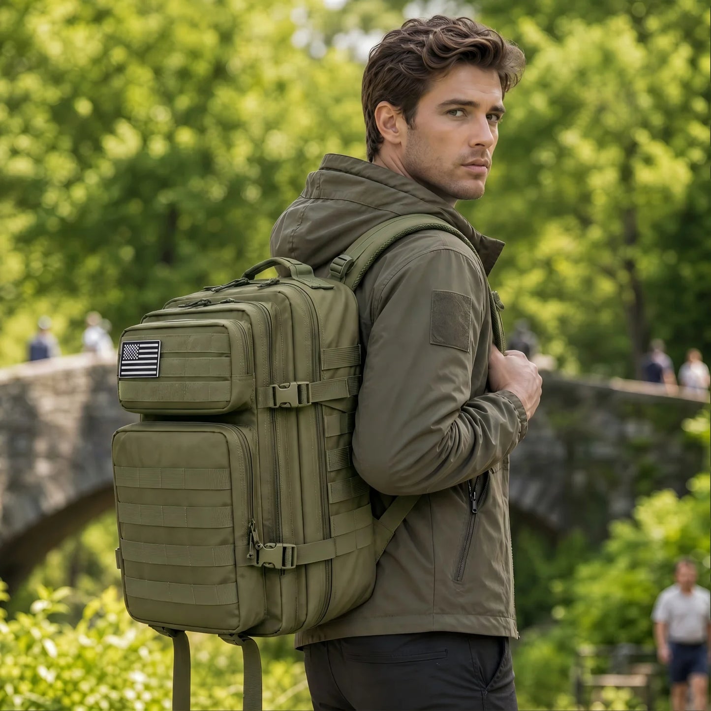 A man wearing an olive green tactical backpack outdoors in a park setting with trees and a stone bridge in the background.