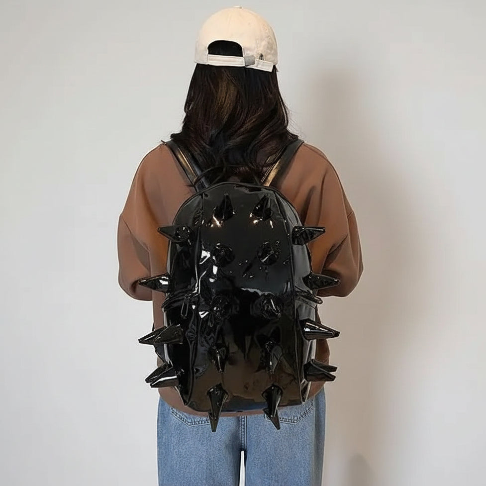 A person viewed from behind wearing a glossy black spiked backpack, a brown sweatshirt, and blue jeans in an indoor setting.
