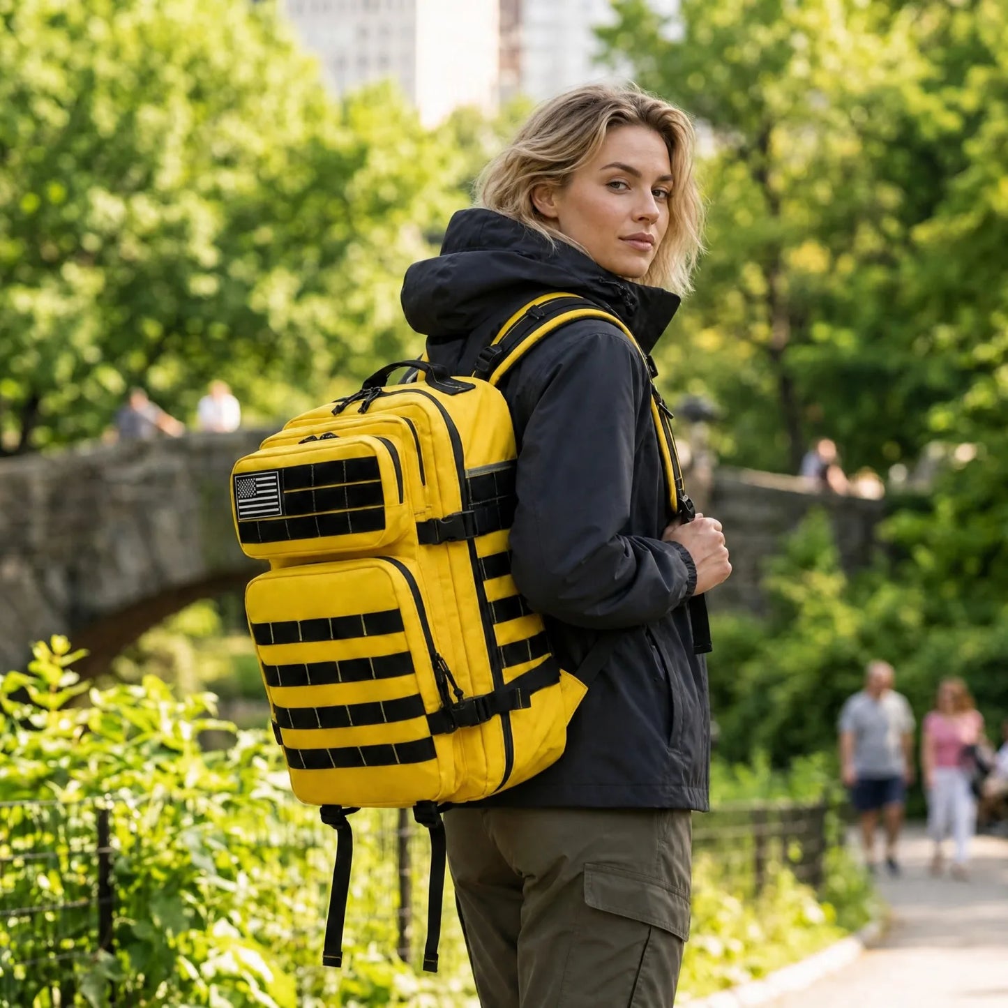 A woman wearing a black hooded jacket and olive cargo pants carries a bright yellow tactical backpack with black webbing and straps in an outdoor park setting.