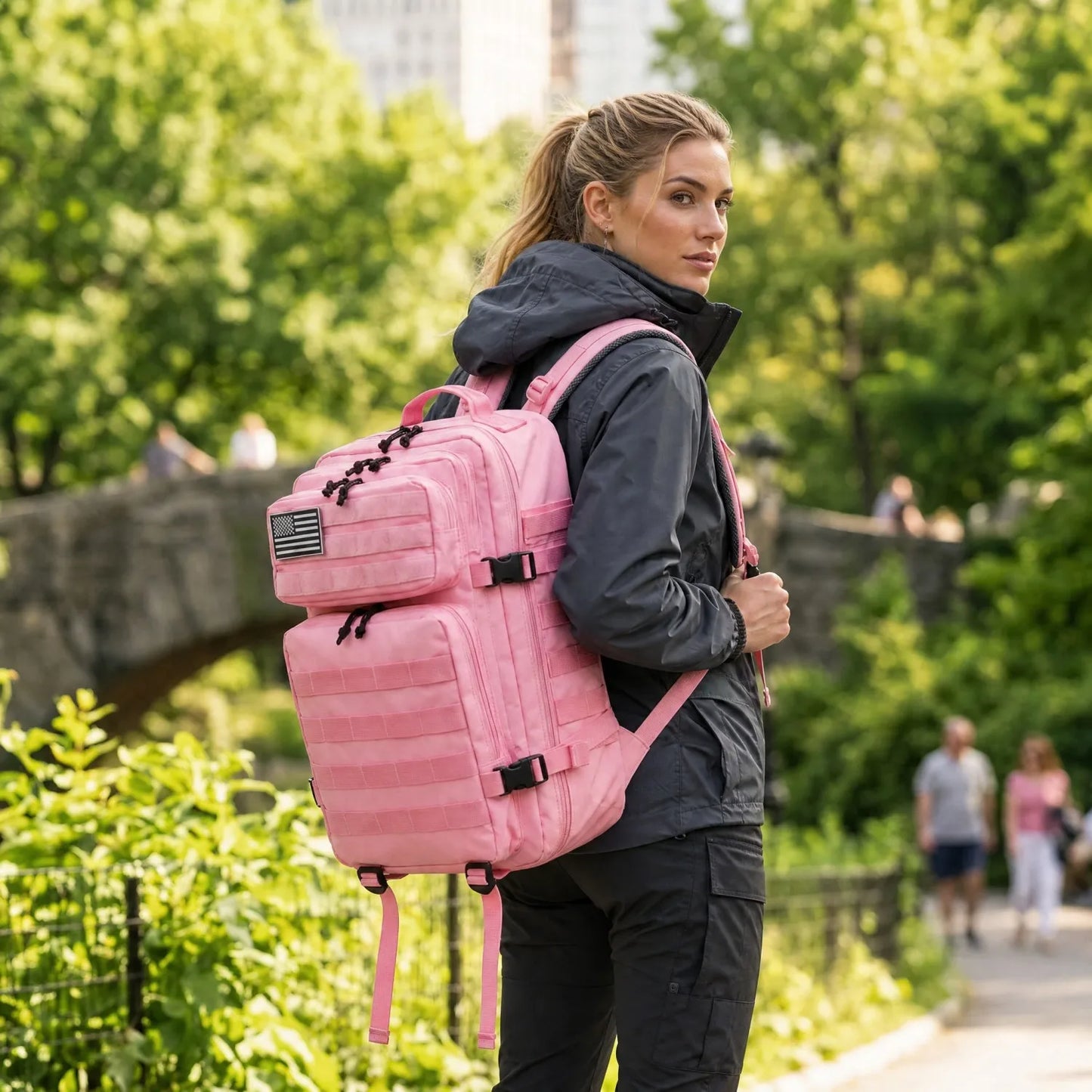 A woman wearing a black jacket and black pants carries a large pink backpack with multiple zippered compartments, photographed outdoors on a park path with trees and blurred pedestrians in the background.