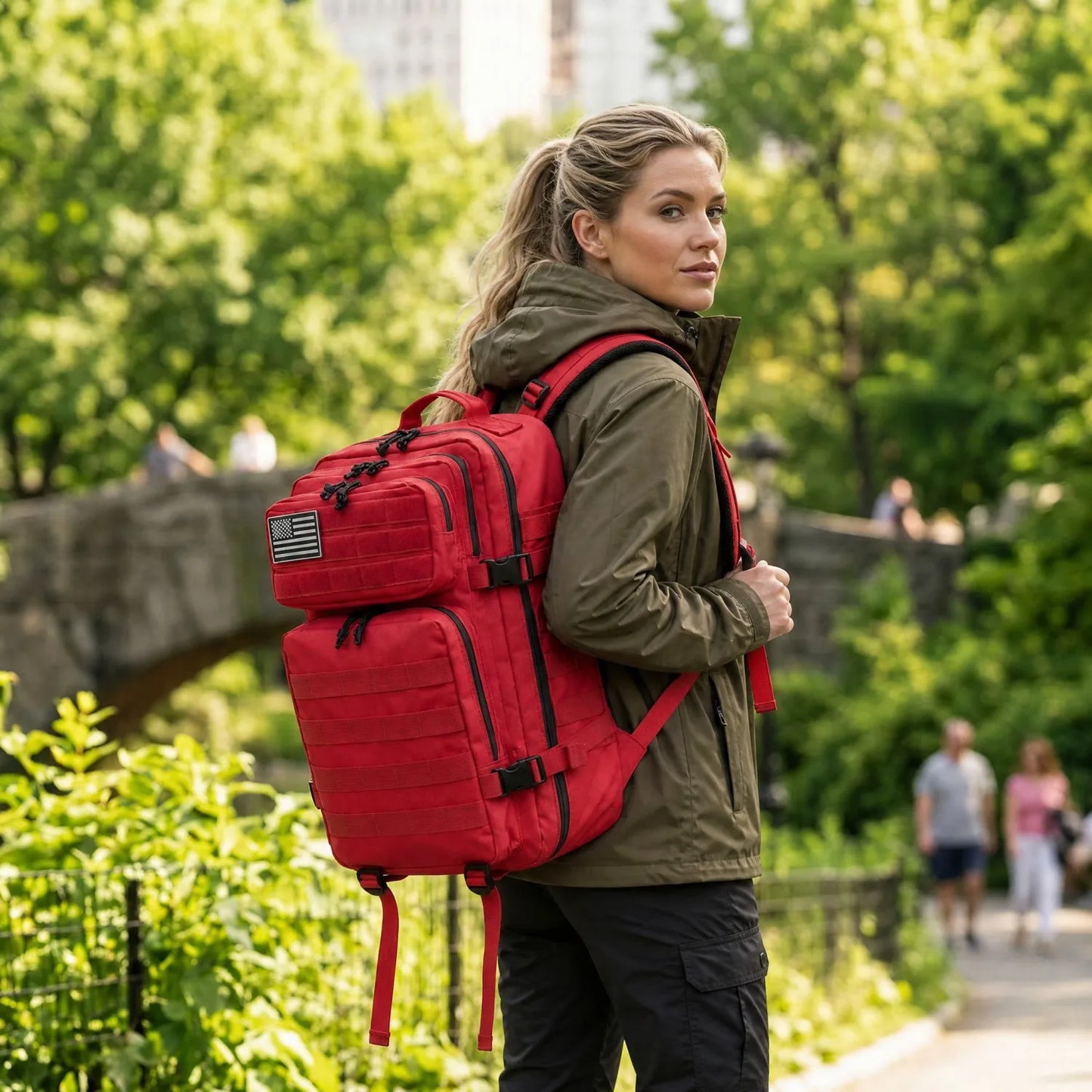 A woman wearing an olive green hooded jacket and black cargo pants carries a large red backpack with black straps and buckles in an outdoor park setting with trees and a stone bridge in the background.