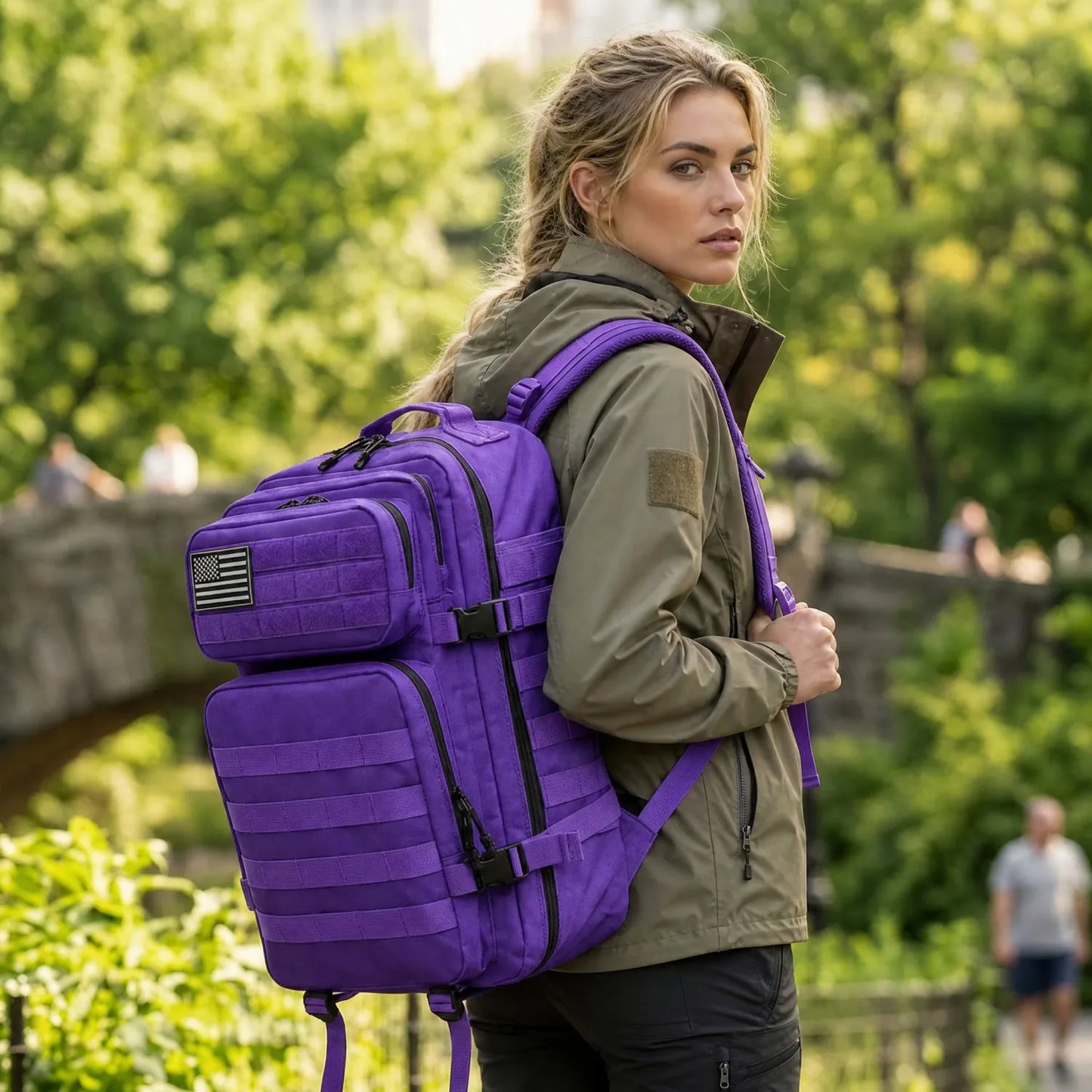 A woman wearing an olive green jacket and black pants is shown from the back in an outdoor park setting, wearing a large purple tactical backpack with black straps and zippers.