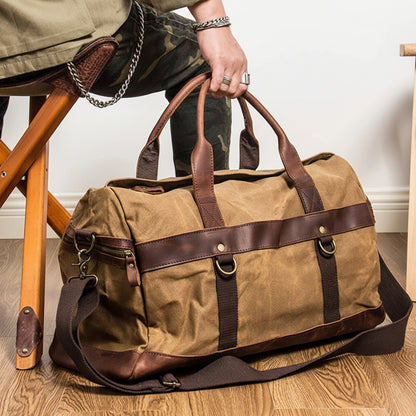 Brown duffel bag with leather straps on a wooden floor next to a person sitting on a chair.