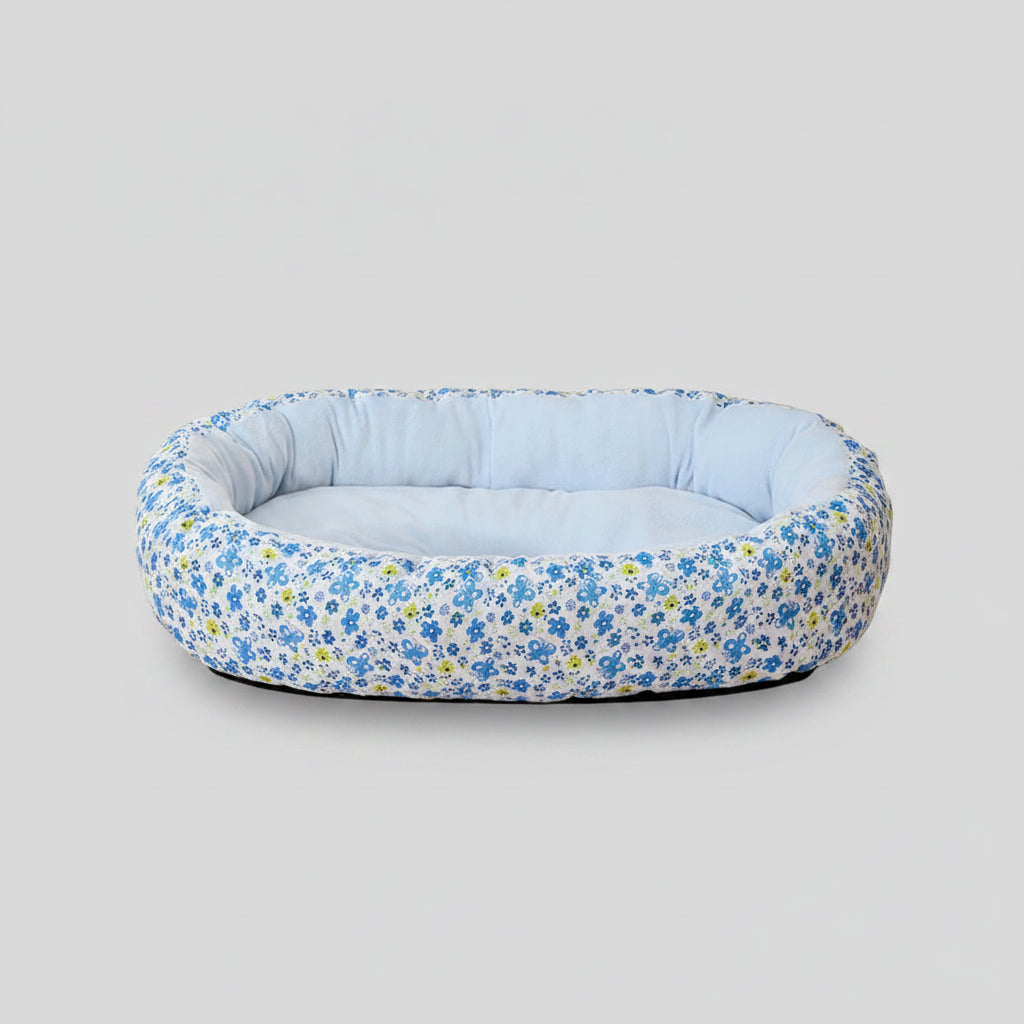 Floral-patterned pet bed on a pink surface with a beige curtain and wooden chair in the background.
