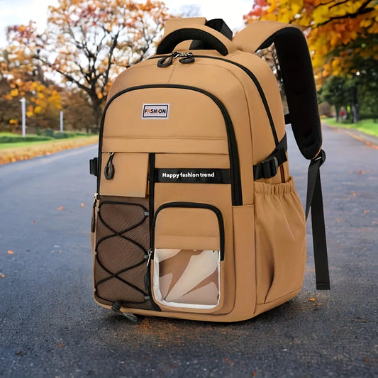 A tan and brown backpack with black straps and trim, placed on an asphalt road with autumn trees in the background. The backpack has multiple front pockets, including a mesh side pocket, a clear zippered pocket