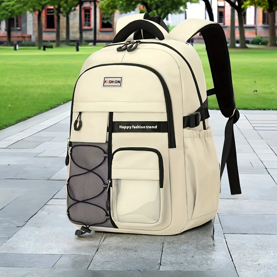 A large beige and black backpack standing on a paved walkway in an outdoor park setting with green grass and trees in the background. The backpack has multiple compartments, a front mesh pocket, side pockets, and a small brand patch that reads 'SHON'.