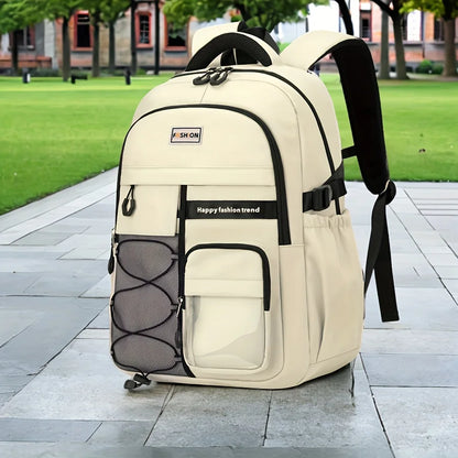A large beige and black backpack standing on a paved walkway in an outdoor park setting with green grass and trees in the background. The backpack has multiple compartments, a front mesh pocket, side pockets, and a small brand patch that reads 'SHON'.